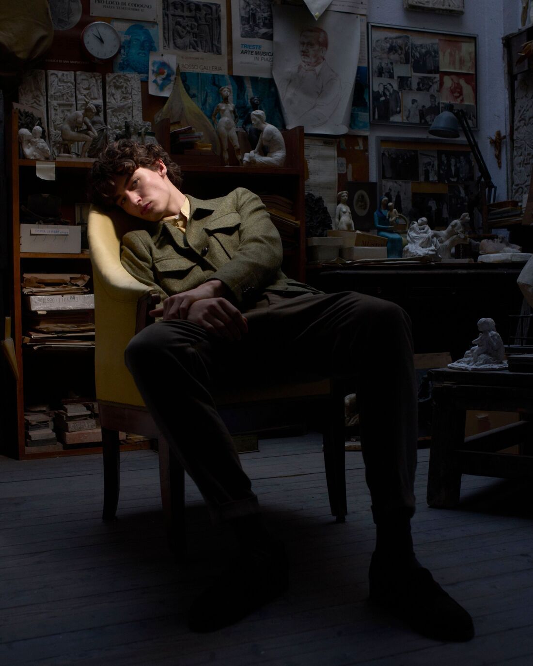 a model sits back relaxed on a chair in a dimly lit room with a soft spotlight on their face, wearing an army green wool jacket